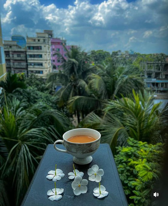 The image features a cup of tea sitting on top of a table, surrounded by white flowers. There are nine flowers in total, with some placed closer to the edge of the table while others are more centrally located. The scene appears to be set outdoors, as there is a cityscape visible through an open window or door nearby.