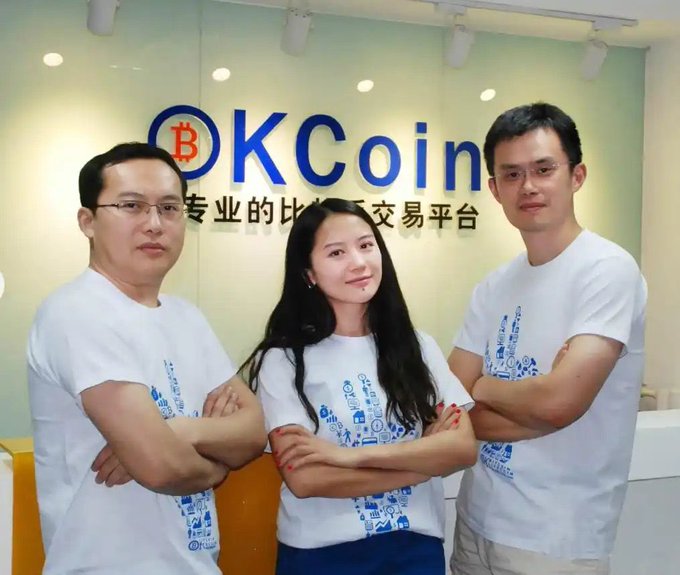 Three people wearing matching white t-shirts with blue designs stand with arms crossed in front of a wall displaying the 'OKCoin' logo and Chinese text reading '专业的比特币交易平台'.