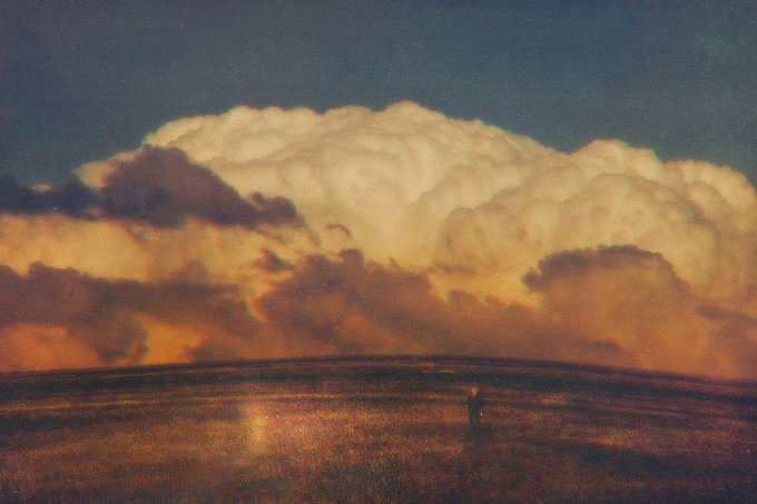A dramatic painting of massive, billowing cumulus clouds illuminated with warm golden light at sunset, rising above a dark, rolling landscape with a small, solitary figure standing in the field.