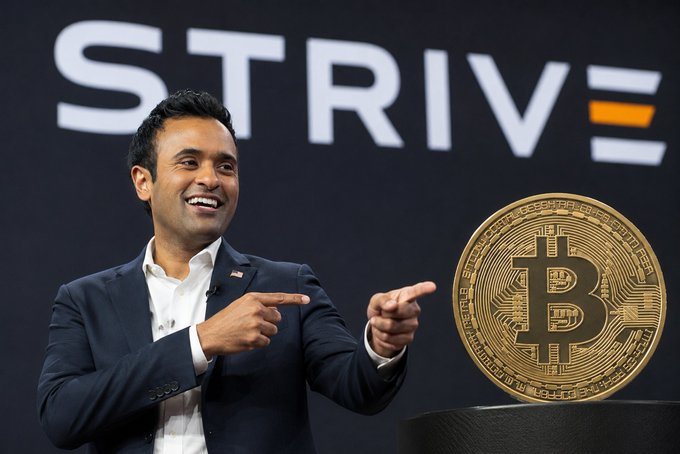 A smiling man in a suit points toward a large Bitcoin display, with the 'STRIVE' logo (with an equal sign) in the background.