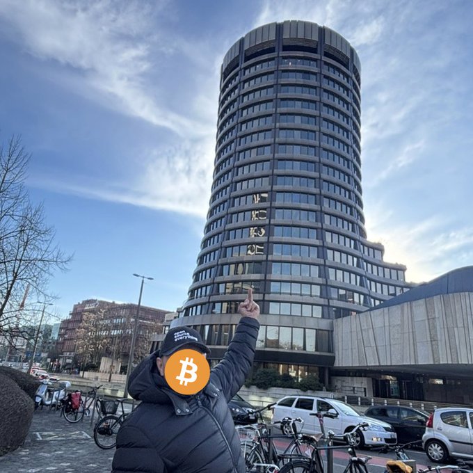 A person points defiantly at the cylindrical BIS tower in Basel, their face obscured by a Bitcoin logo, symbolizing the rejection of the central banking cartel in favor of decentralized monetary sovereignty.