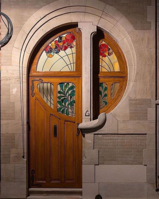 An ornate wooden door with stained glass floral panels set into an arched stone entryway, featuring decorative Art Nouveau-style metalwork.
