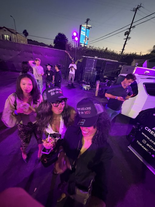 A group of young women wearing 'ROOF LIKE A DOG ON MTU' trucker hats pose for a selfie at a dusk outdoor gathering near a vehicle, with purple lighting illuminating the scene.