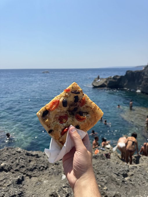 A hand holds up a rectangular slice of focaccia topped with tomatoes and olives, with a rocky beach, blue water, and swimmers visible in the background.