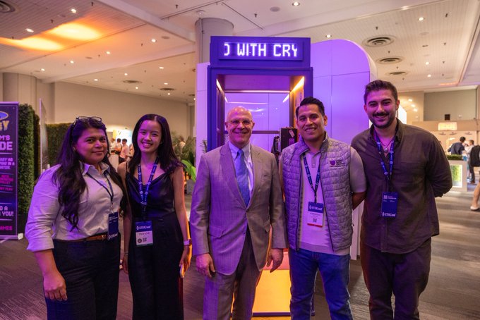 The image shows a group of five people posing for a photo at an event. They are standing next to each other, with one person wearing a suit and tie while another has a name tag around their neck. The backdrop features a sign that reads "J WITH CRY", which is likely related to the event or cause they are promoting. A man in a suit stands out among them, possibly representing SWC or another organization involved in the event.