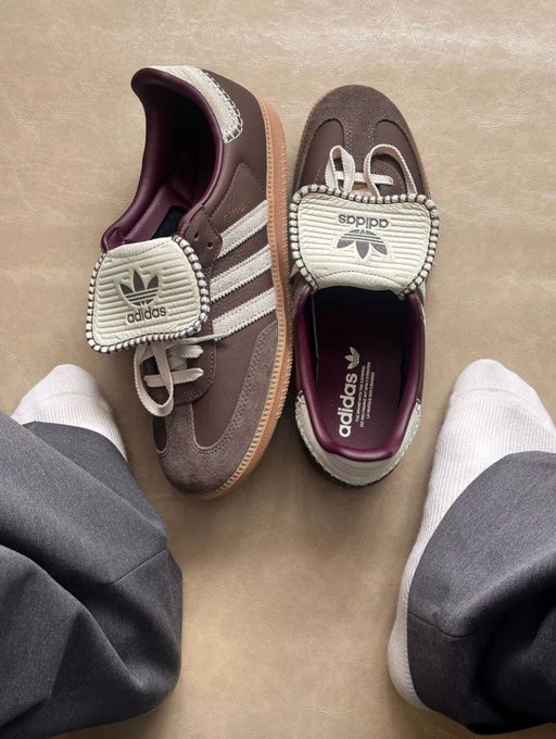 A pair of brown Adidas Samba sneakers with cream-colored tongue tags featuring the Adidas trefoil logo and three-stripe detailing, photographed from above on a beige surface next to someone wearing white socks and gray pants.