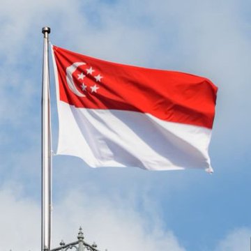 The image shows the Singapore flag waving against a partly cloudy sky, featuring its distinctive red and white horizontal bands with a white crescent moon and five stars in the upper left corner. This flag visually represents Singapore, the home country of Genius Group, the company mentioned in the post that plans to purchase $827 million worth of Bitcoin over the next five years.