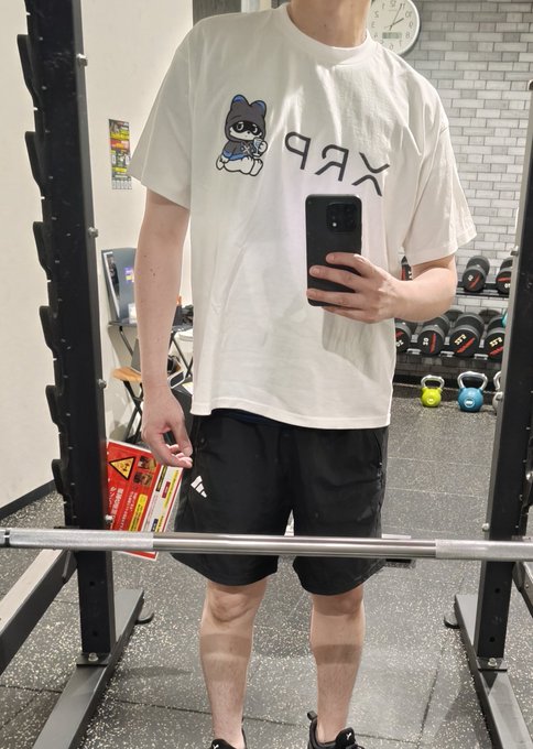 A mirror selfie of a person wearing a white t-shirt with an upside-down 'XRP' logo and cat graphic, black shorts, standing in a gym.