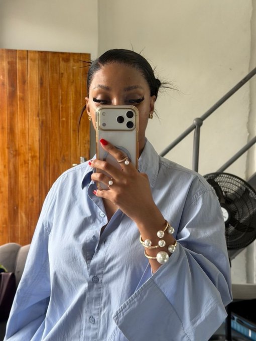 A mirror selfie of a woman wearing an oversized light blue button-up shirt, holding an iPhone with a clear case, adorned with gold jewelry and pearl bracelets, with red nail polish.