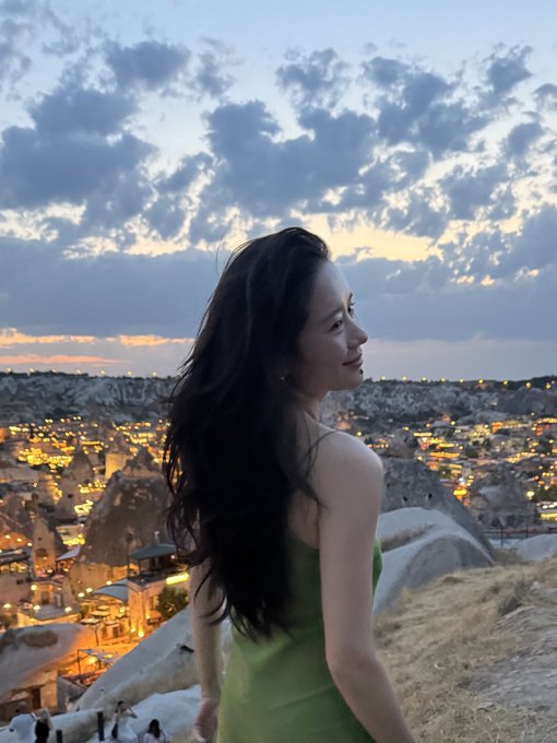 A woman with long dark hair in a green dress looks out over a scenic valley with illuminated rock formations and a dusky sky. The image appears to capture a travel moment, possibly from Cappadocia, Turkey, rather than directly relating to crypto or trading platforms.