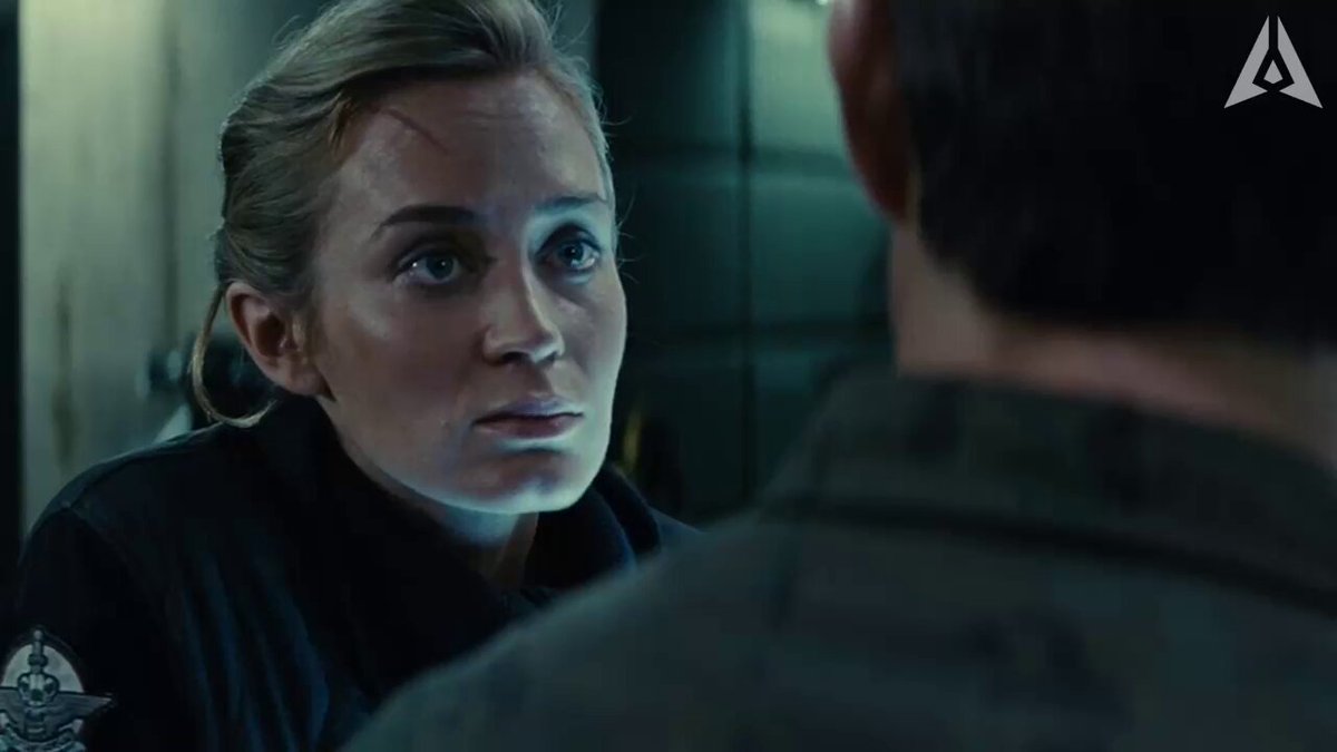 The image shows a woman wearing a black jacket standing next to a man who is also dressed in black. They are both looking at each other, possibly having a conversation or discussing something important. The scene takes place indoors with a green wall as the background.