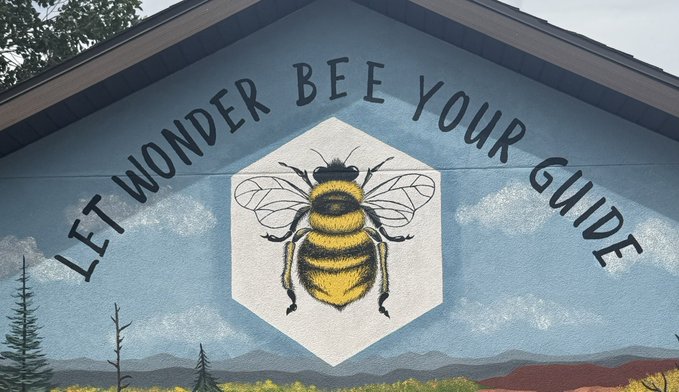 A mural painted on a blue wall depicts a yellow and black bumblebee inside a white hexagon, with the text 'LET WONDER BEE YOUR GUIDE' arcing above it, set against a backdrop of painted clouds and landscape.