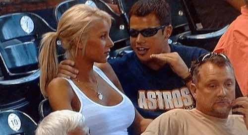 A photo of a young blonde woman in a white tank top sitting next to a man wearing an Astros shirt and sunglasses in what appears to be stadium seating, with another man visible in the foreground.