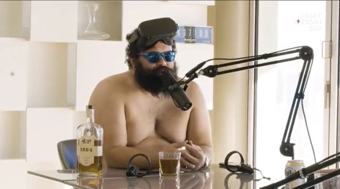 A shirtless, bearded man wearing sunglasses and a VR headset sits at a podcast microphone with a glass and bottle of "1884" bourbon whiskey, illustrating the bizarre premise behind the caption "then how do you explain this?"
