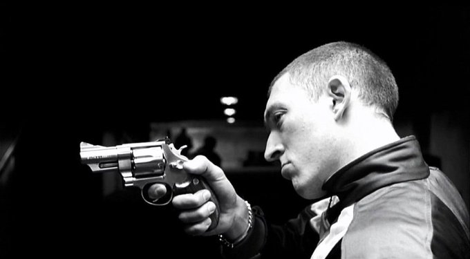 A black and white profile photo of a young man with short hair aiming a silver revolver in a dimly lit setting.