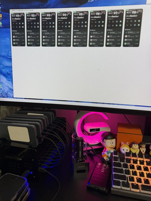 A desk setup featuring a monitor displaying eight "Seeker" smartphone mockups numbered 01-08, accompanied by a multi-device charging station with several phones connected, alongside a keyboard with figurines. The image directly ties to the post by visually emphasizing the user's collection of Seeker phones and deep involvement in the Solana/World ecosystem, making a case for the airdrop they're hoping for from @world_xyz.