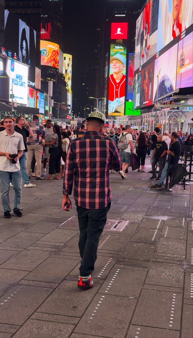 A man in a red and black plaid shirt and dark pants walking through a crowded, brightly lit Times Square at night.