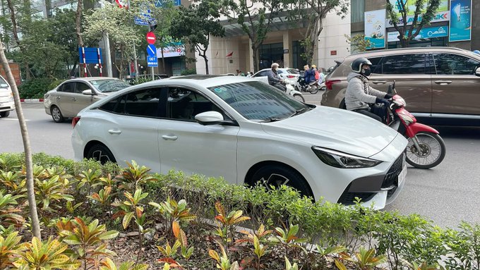 A white sedan parked next to a hedge on a city street, with motorbikes and other vehicles visible in the background of an urban setting.