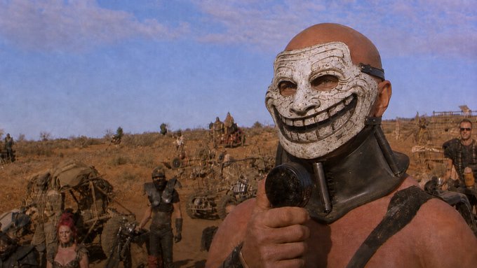 A scene from a post-apocalyptic film showing a bald man wearing a grotesque smiling mask and a metal neck brace, set against a dusty desert landscape with vehicles and other characters in the background.