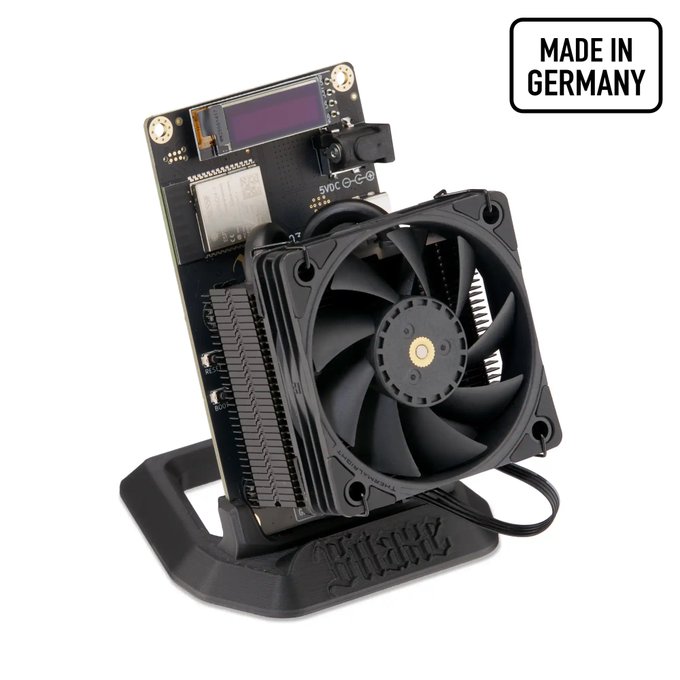 A Bitaxe Bitcoin mining device featuring a circuit board with display screen and large black cooling fan mounted on a 3D-printed stand, labeled 'MADE IN GERMANY' in the corner.