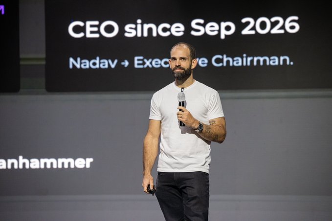A man in a white t-shirt holds a microphone on stage in front of a screen reading 'CEO since Sep 2026, Nadav → Executive Chairman.'