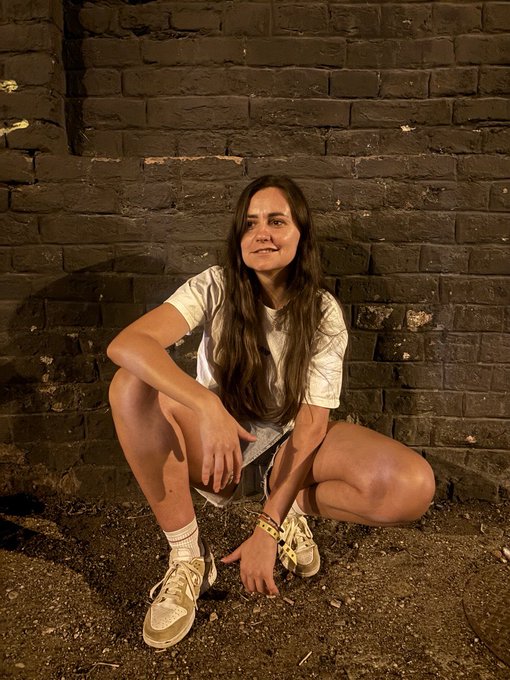 A woman with long brown hair crouches against a dark brick wall, wearing a white t-shirt and light sneakers, with a yellow wristband visible on her wrist.