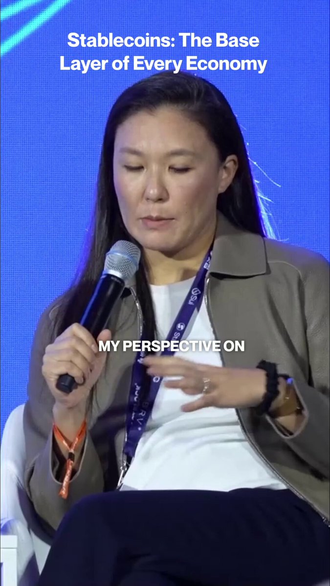 The image shows a woman sitting down with her hands clasped together while holding a microphone. She appears to be speaking or addressing an audience at an event. The background of the image features a blue wall, which contrasts with the brown jacket she is wearing. There are also orange wristbands visible on her wrists.