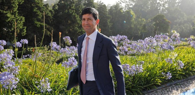 A man in a dark suit and pink tie stands confidently in a garden filled with purple agapanthus flowers, smiling at the camera during golden-hour sunlight. The image appears unrelated to the post's content about crypto trading and the weekly macroeconomic calendar of events like PMI releases and FOMC minutes.