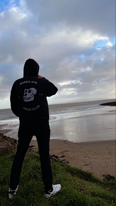 A person wearing a black 'Bored Ape Yacht Club' hoodie stands on a grassy cliff overlooking a beach and ocean under a cloudy sky.