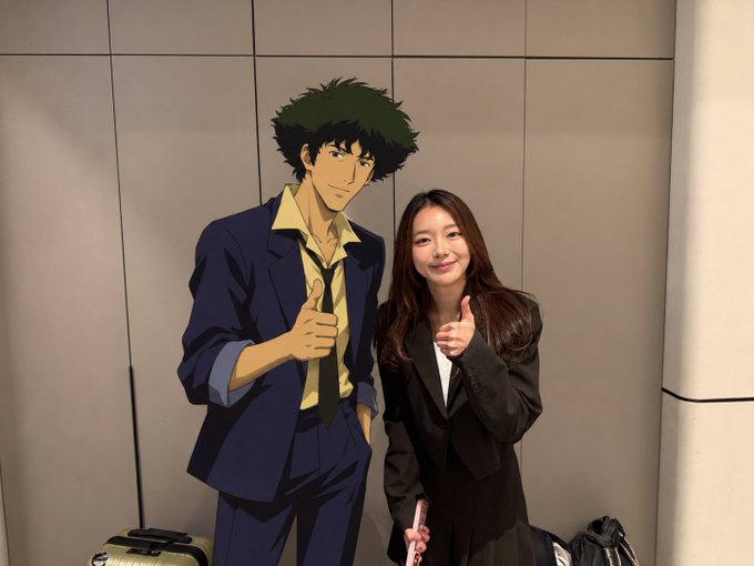 A woman in a black suit poses next to a life-sized cardboard cutout of the anime character Spike Spiegel from Cowboy Bebop, both giving thumbs up.