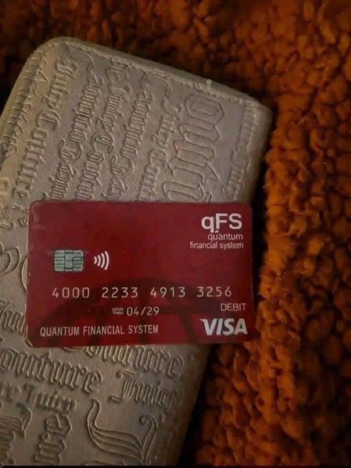 A red debit Visa card for 'qFS quantum financial system' with the number 4000 2233 4913 3256 resting on a textured beige wallet or fabric.