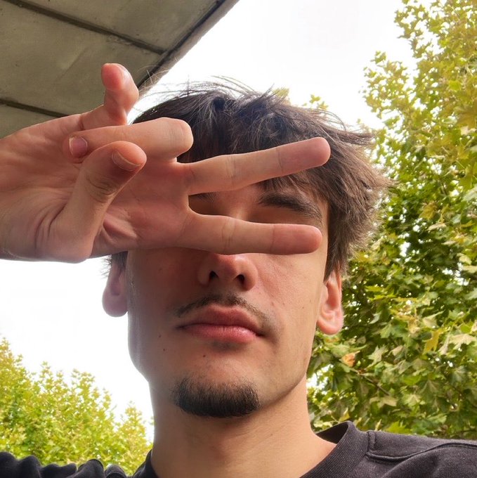 A young man with messy brown hair and a goatee holds up a peace sign over his eye, standing outdoors against a backdrop of green trees and a bright sky. The "YO GANG" caption matches his casual, laid-back selfie vibe as he flashes a chilled-out greeting to his followers.