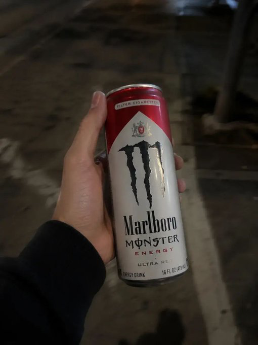 Hand holding a red and silver can of 'Marlboro Monster Energy Ultra Red' energy drink (16 fl oz / 473 mL), photographed on a dimly lit tiled sidewalk.