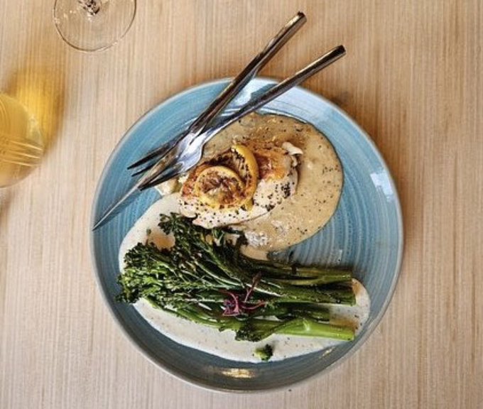 An overhead view of a blue plate containing grilled fish, lemon, broccolini, and creamy sauce, with a fork and knife and a glass of white wine beside it.