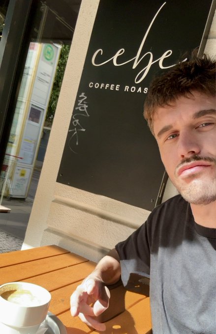 A selfie of a young man with a mustache sitting at an outdoor café table with a coffee, in front of a storefront sign reading 'cebe COFFEE ROAS[TERS]'.