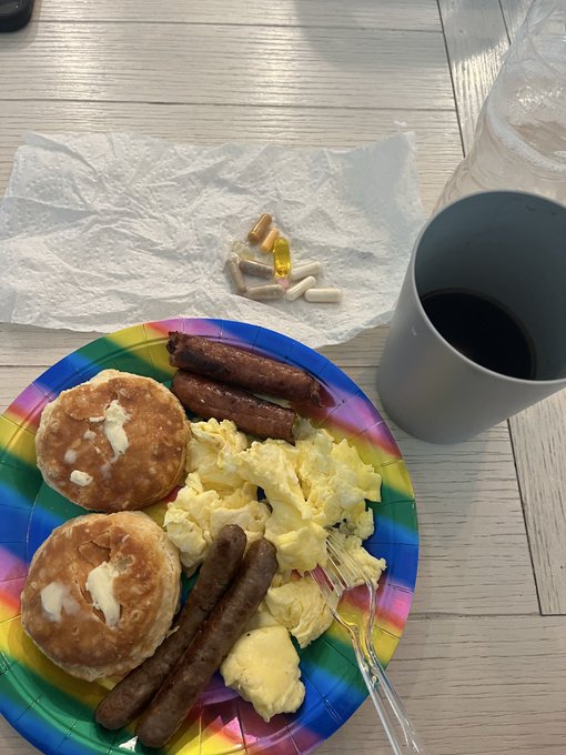 A breakfast plate with pancakes, sausage links, and scrambled eggs on a rainbow-colored plate, accompanied by a mug and a napkin with pills and capsules.