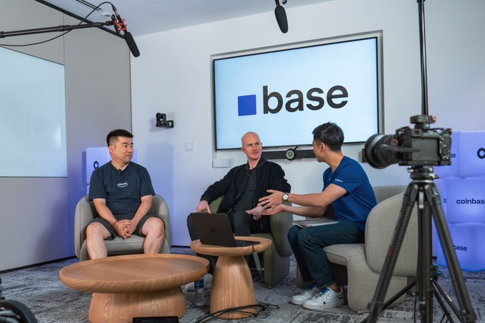 Three men sit in a studio setting with a screen displaying the 'base' logo behind them, with filming equipment including microphones, a camera on a tripod, and lights visible in the room.