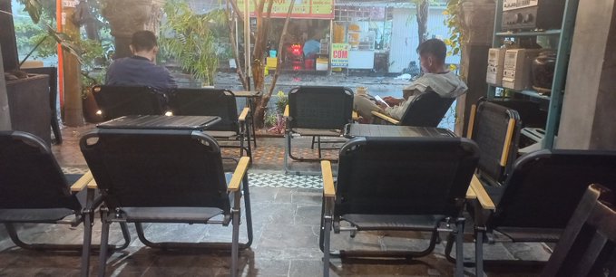 Two people sitting in black chairs on a patio looking at their phones, with a laptop on a chair in the foreground and a street visible through the entrance.
