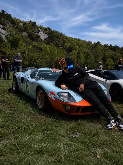 A person wearing a beanie and black "ENÒ" sweatshirt leans against a vintage Gulf-liveried race car (#6) at an outdoor car gathering under a blue sky. The motivational message about seizing the day pairs with the image's theme of ambition and drive, using the classic racing scene as a visual metaphor for perseverance and progress.