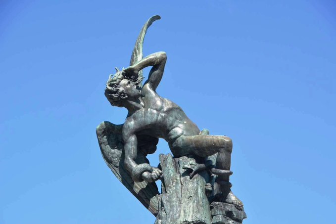 A weathered bronze statue of a winged figure—likely the archangel Michael vanquishing Satan—stands defiantly atop a pedestal against a clear blue sky, its sword raised in a striking pose. The image visually mirrors the post's warning about "crossing a constitutional red line," evoking themes of moral struggle, defiance, and the violation of a sacred boundary.