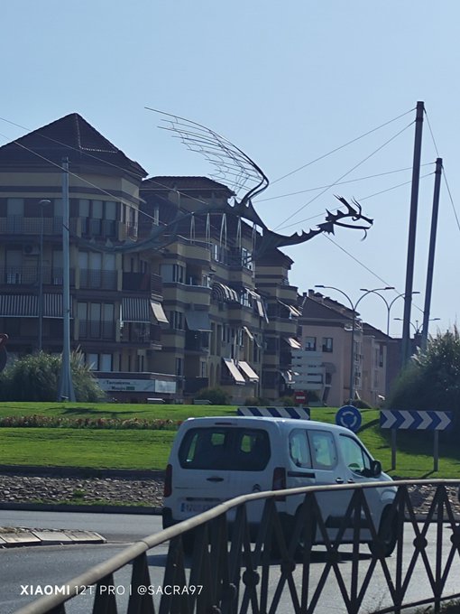 A street scene with apartment buildings, a white car, and a striking metal sculpture of a deer silhouette with antlers made of curved rods, taken with a Xiaomi 12T Pro.
