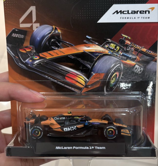 A hand holds a packaged McLaren Formula 1 die-cast toy car (#4, featuring OKX, Dell, Chrome, and Workday sponsorships) in front of its blister-pack backing card, which displays the "McLaren Formula 1 Team" logo and an image of the race car. The post playfully teases that for now this is just a toy, but hopes one day they'll get to see the real McLaren F1 car up close, with the OKX branding prominently featured tying into their sponsorship partnership.