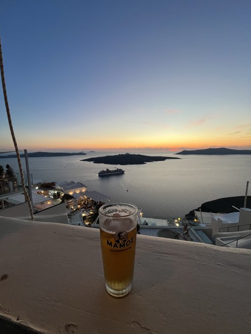 A glass of beer from MAMOS brewery sits on a white ledge overlooking a stunning sunset over the Aegean Sea, likely Santorini, with a cruise ship visible in the distance and white cliffside buildings below.