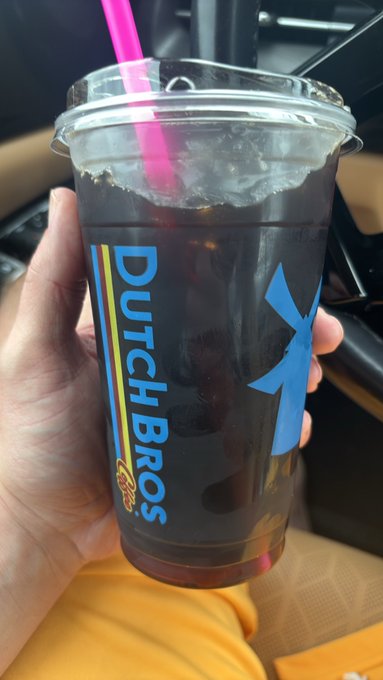 A hand holds a clear plastic Dutch Bros cup filled with a dark beverage and ice, with a pink straw, taken inside a vehicle.