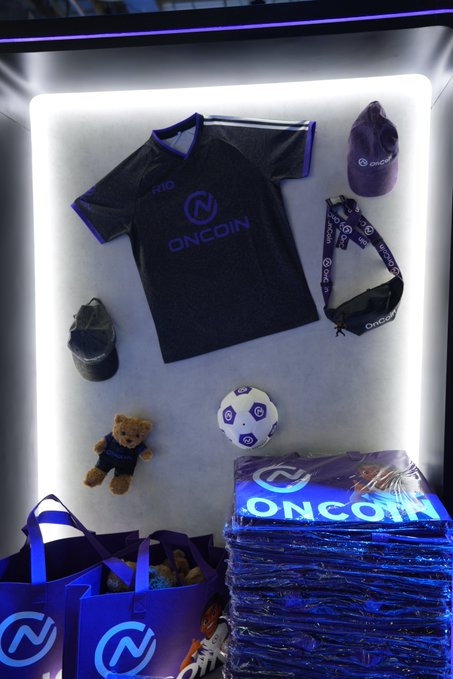 The image showcases a brightly lit display at the **OnCoin** booth at TOKEN2049, featuring branded merchandise including a black "ONCOIN" t-shirt, a cap, lanyards, a teddy bear, a soccer ball, and stacks of promotional tote bags and drawstring sacks with the OnCoin logo, attracting a lively crowd of attendees and familiar faces.