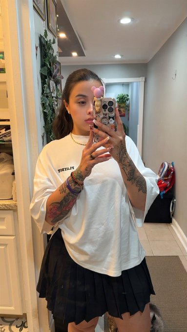 A young woman with arm tattoos takes a mirror selfie in a bathroom or hallway, wearing a white t-shirt, black pleated skirt, and a pink heart filter over one eye.