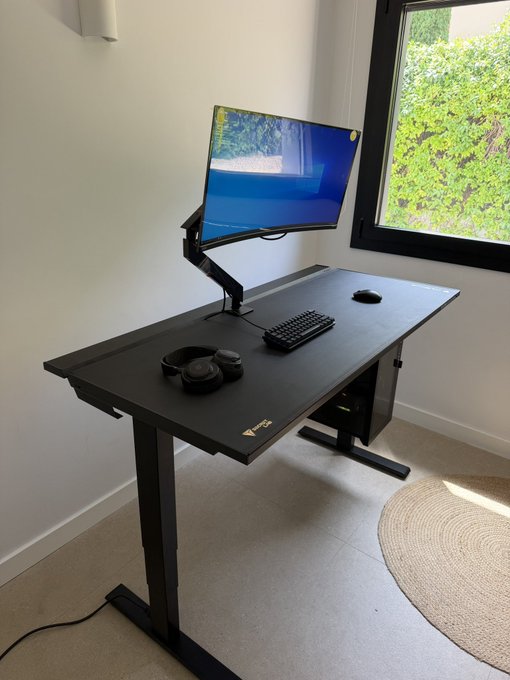 A minimalist home office setup featuring a black standing desk with an ultrawide curved monitor mounted on an adjustable arm, a compact mechanical keyboard, mouse, and headphones, positioned near a window overlooking greenery. The clean, distraction-free workspace reflects the post's message of focus and productivity, suggesting the trader is ready to capitalize on a 100 SOL gain.