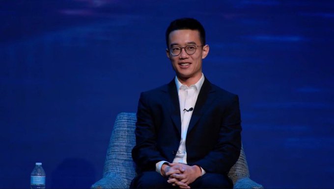 A man in a dark suit and glasses sits smiling on a stage with a blue background, with a small water bottle visible beside him.