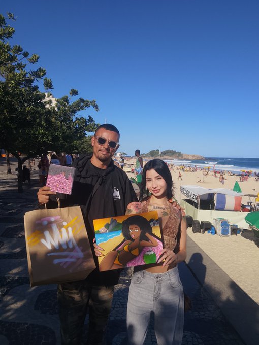 A man and woman pose on a sunny beach boardwalk, holding up artwork and a shopping bag, with beach umbrellas and the ocean behind them.