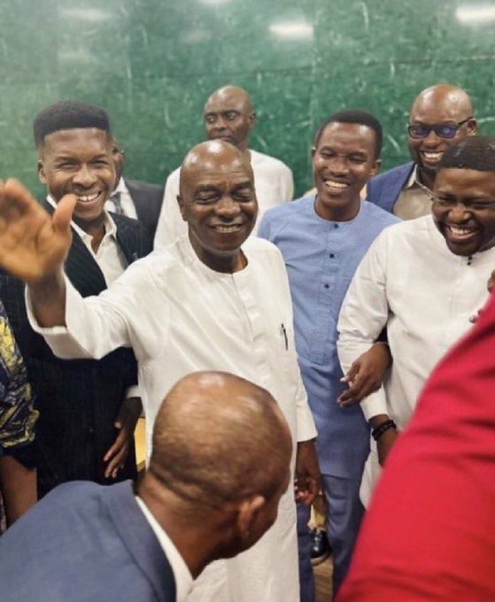 A joyful group of men in formal attire share laughter and smiles in a warmly lit indoor setting, with the central figure waving warmly to the crowd, capturing the celebratory spirit fitting for an upcoming $INJ summit in Washington DC on July 16th.
